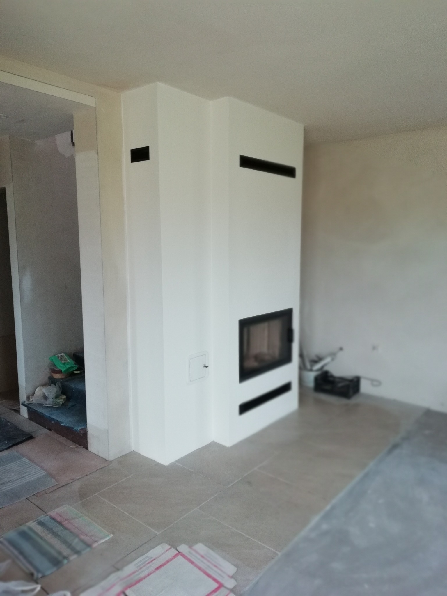 Biały, nowoczesny kominek z czarnym wkładem, w trakcie wykańczania wnętrza. Widoczne narzędzia i materiały budowlane. Minimalistyczny design.