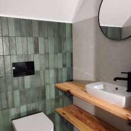 Tomasz Górski - Nowoczesna łazienka z zielonymi, pionowo ułożonymi płytkami, wiszącą toaletą, drewnianym blatem pod umywalką i okrągłym lustrem.