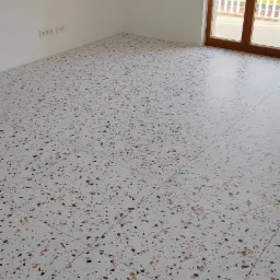 Nowoczesna posadzka z płytek lastryko w jasnym wnętrzu, widok na ścianę z gniazdkami i okno balkonowe z widokiem na budynek.