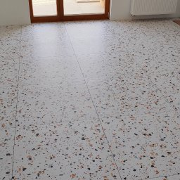 Tomasz Górski - Biała posadzka terazzo z kolorowymi, drobnymi kamyczkami, widoczna z perspektywy podłogi, z oknem balkonowym i grzejnikiem w tle.