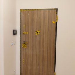 Tomasz Górski - Nowe drzwi wewnętrzne z okleiną drewnopodobną, zabezpieczone folią i taśmą malarską podczas prac wykończeniowych, widoczny fragment podłogi z lastryko i klimatyzator.