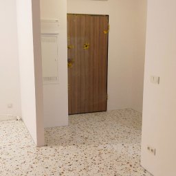 Tomasz Górski - Nowoczesne wnętrze z terazzo na podłodze, drewniane drzwi zabezpieczone folią ochronną, białe ściany i klimatyzacja na górze.