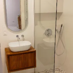 Nowoczesna łazienka z umywalką nablatową na drewnianej szafce, kabina prysznicowa typu walk-in z deszczownicą i odpływem liniowym oraz podłoga z lastryko.