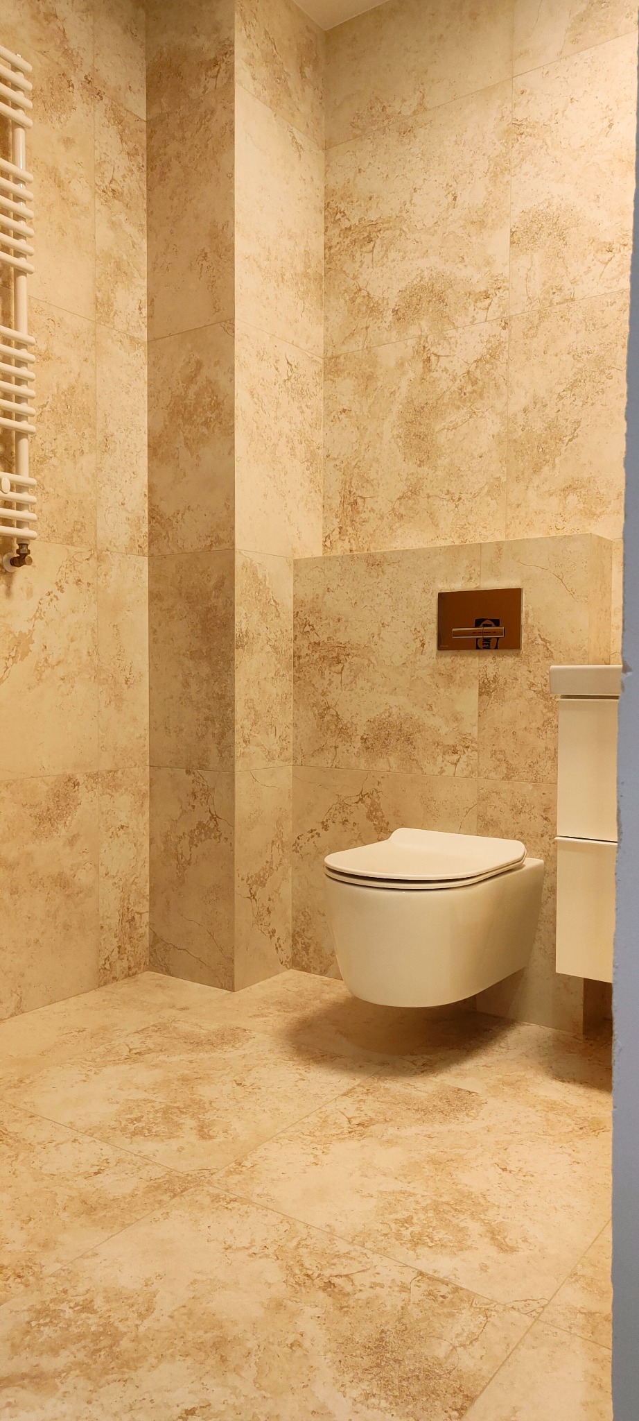 Beżowa łazienka z wiszącą toaletą, grzejnikiem drabinkowym i płytkami imitującymi marmur na ścianach i podłodze.