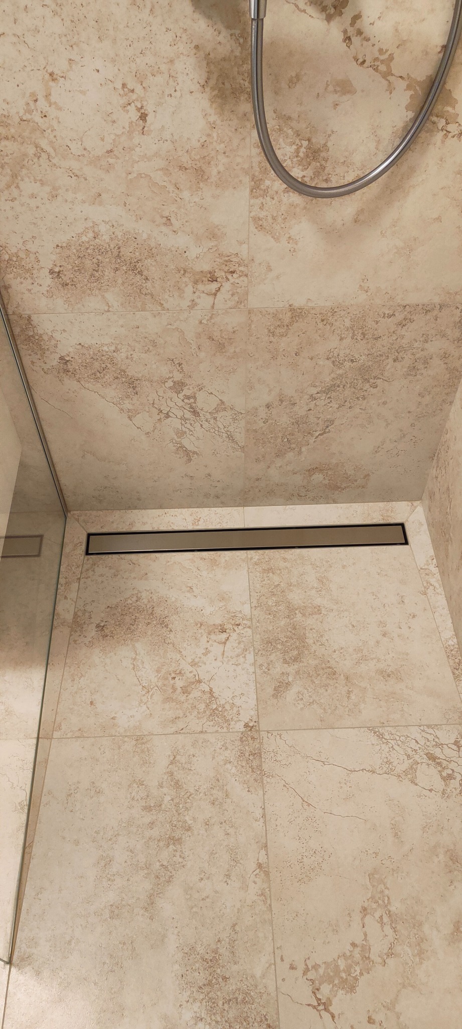 Beżowe płytki imitujące trawertyn w kabinie prysznicowej z odpływem liniowym i szklaną ścianką.