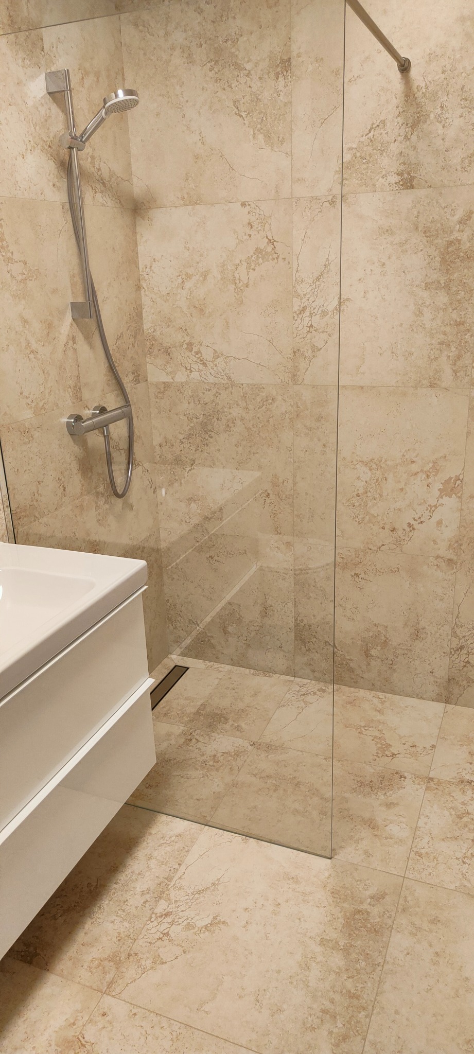 Nowoczesna kabina prysznicowa ze szklaną ścianką i beżowymi płytkami imitującymi marmur, widoczny odpływ liniowy w podłodze i biała szafka pod umywalką.