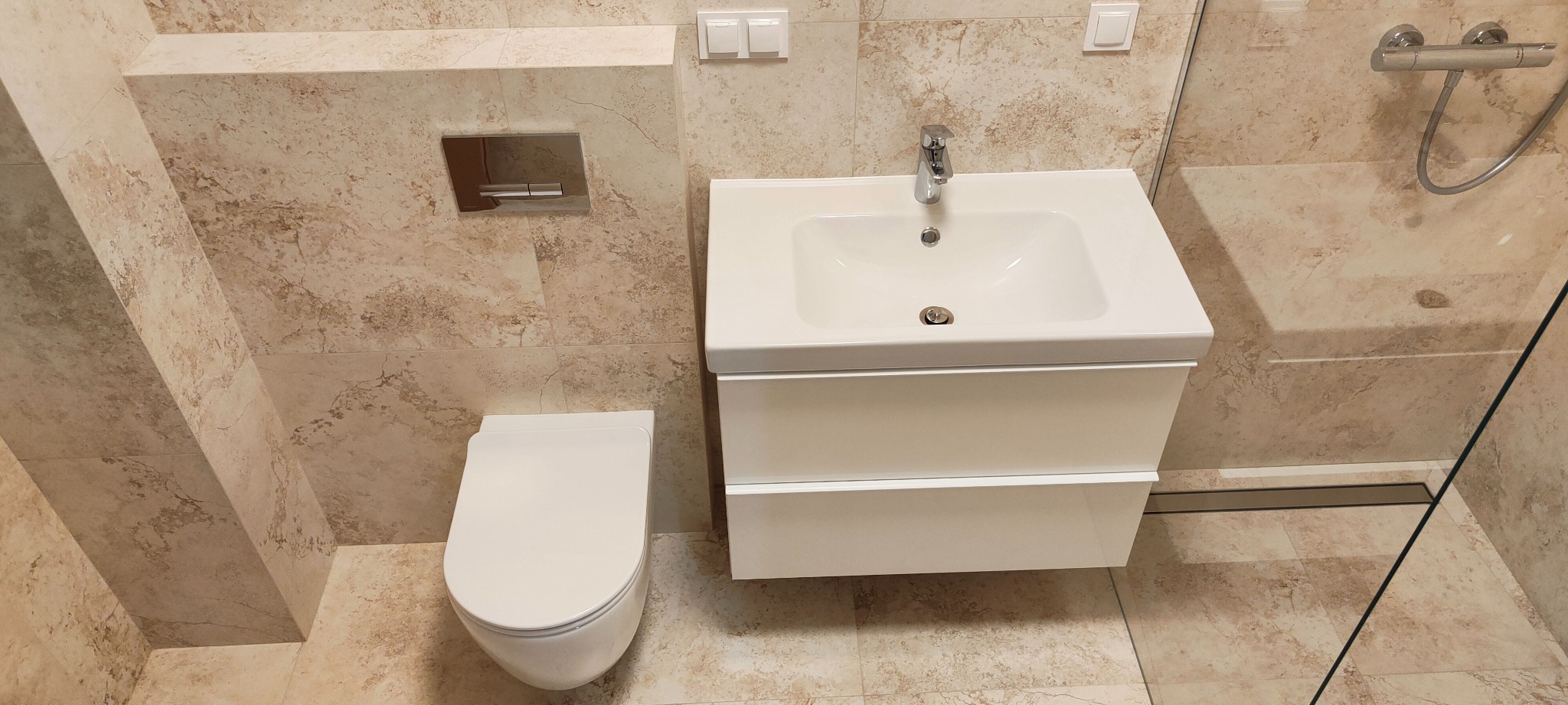 Nowoczesna łazienka z beżowymi płytkami imitującymi marmur, białą wiszącą miską WC, umywalką z szafką i kabiną prysznicową ze szklaną ścianką.