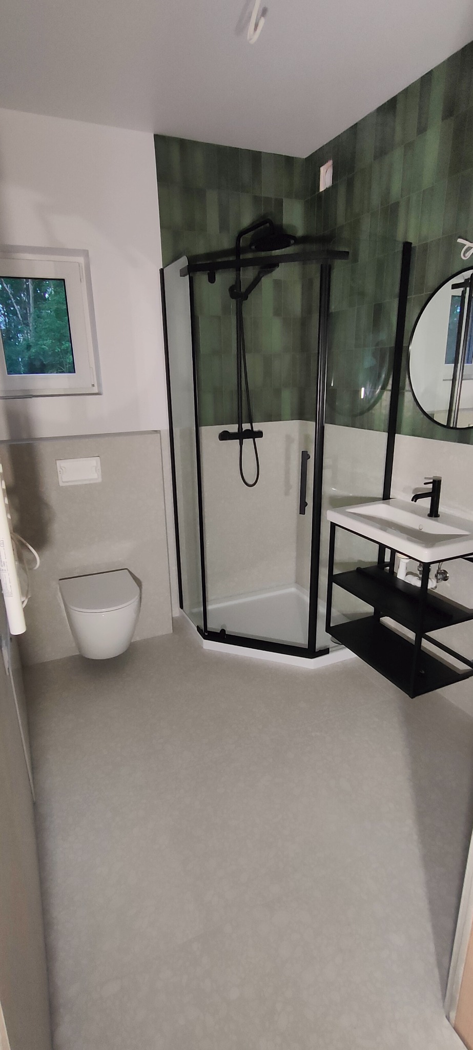 Nowoczesna łazienka z kabiną prysznicową z czarnymi ramami, umywalką na czarnym stelażu, toaletą podwieszaną i zielonymi płytkami na ścianie.