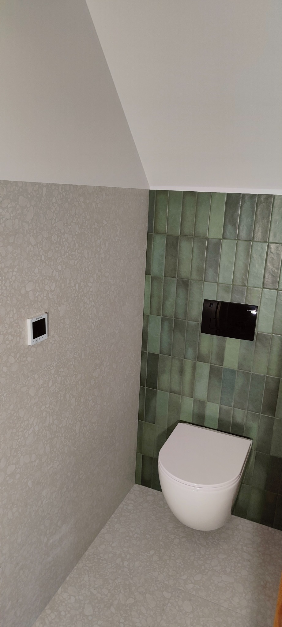 Wykończona łazienka w Żyrardowie z wiszącą toaletą na tle zielonej glazury i ściany z szarą mozaiką, widoczny również panel sterowania ogrzewaniem.