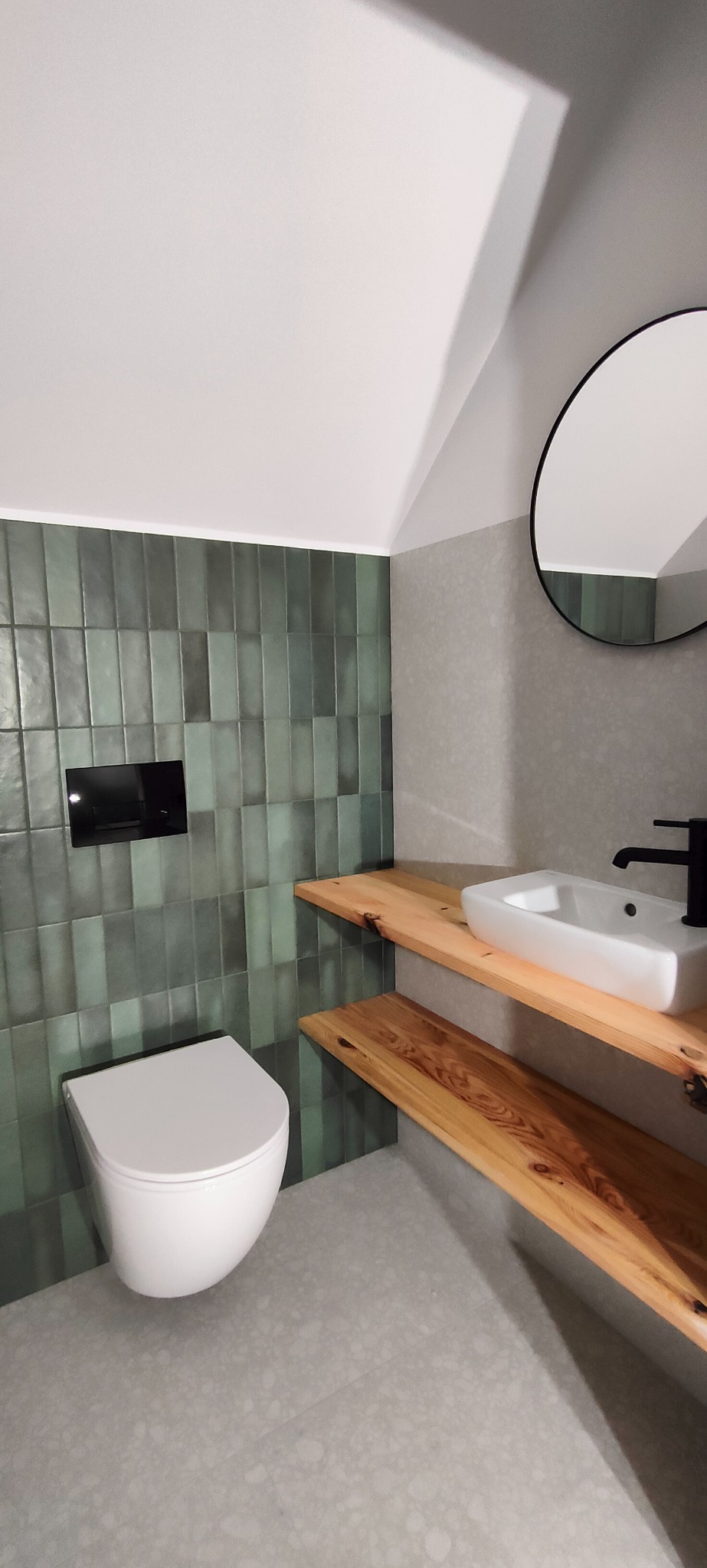 Nowoczesna łazienka z zielonymi, pionowo ułożonymi płytkami, wiszącą toaletą, drewnianym blatem pod umywalką i okrągłym lustrem.