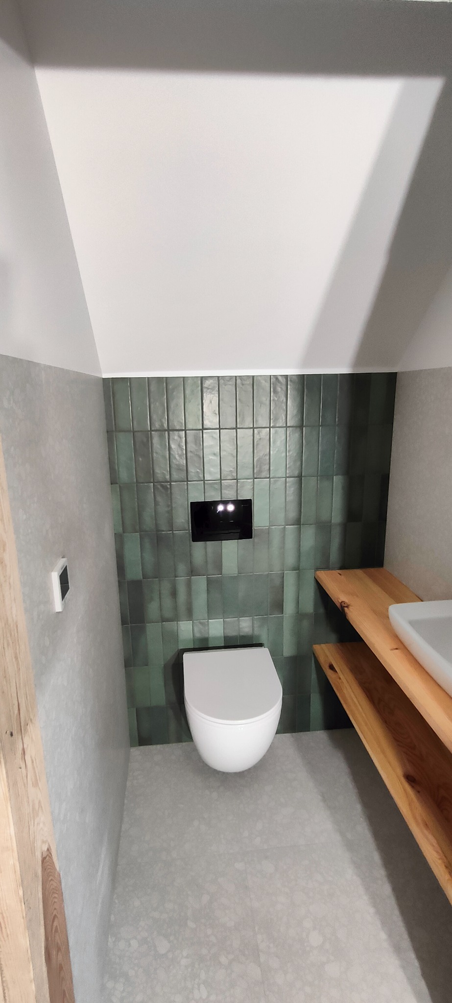 Nowoczesna łazienka na poddaszu z zielonymi, pionowo ułożonymi płytkami, podwieszaną toaletą i umywalką na drewnianym blacie.