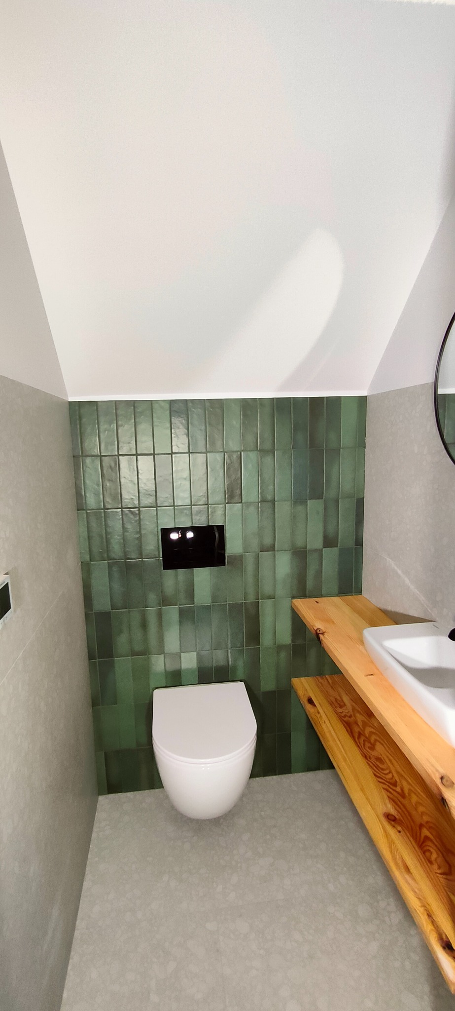 Nowoczesna łazienka w odcieniach szarości i zieleni z wiszącą toaletą i drewnianą umywalką na tle ściany z zielonej, pionowo ułożonej glazury.