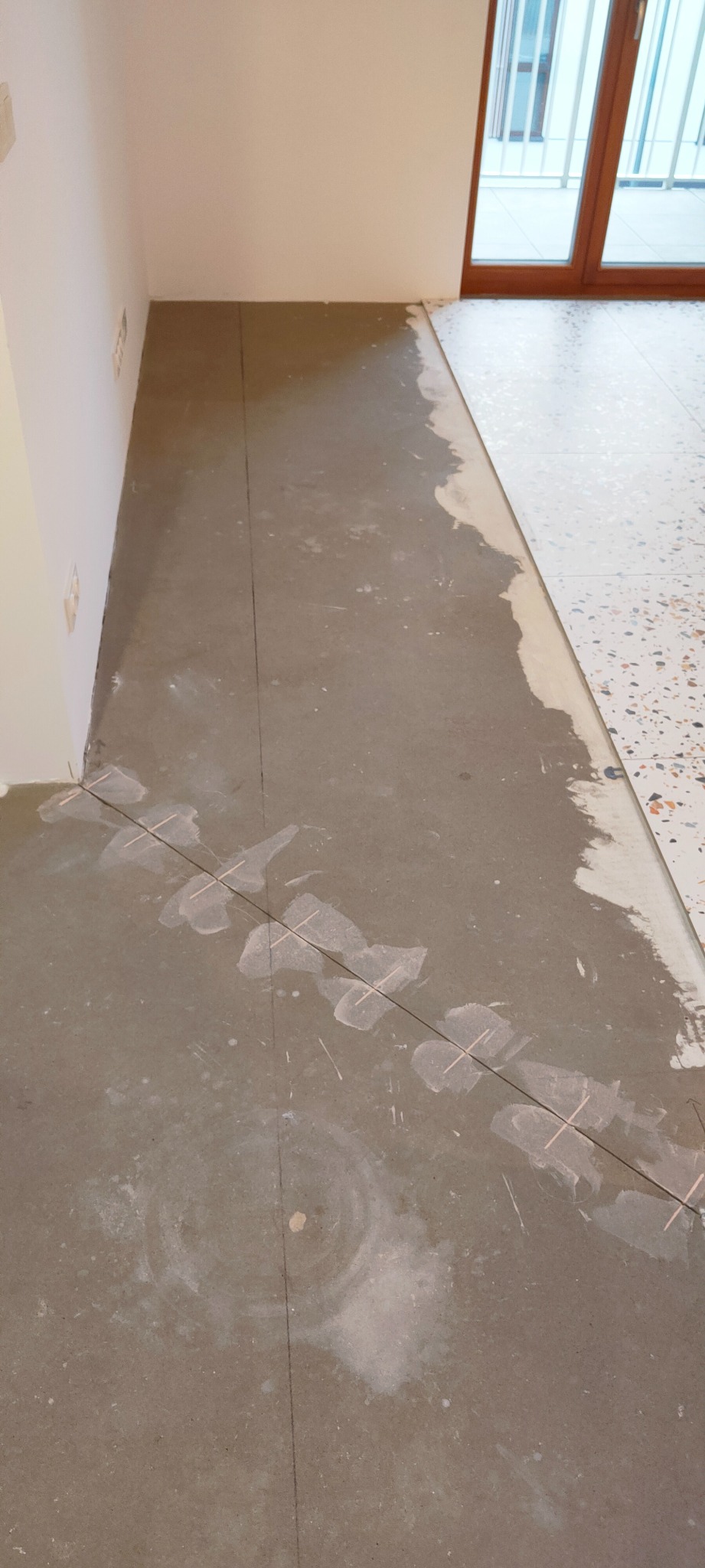 Podłoga w trakcie układania płytek, widoczne sznurki i krzyżyki dystansowe, częściowo ułożone płytki lastryko, surowy beton pod spodem.