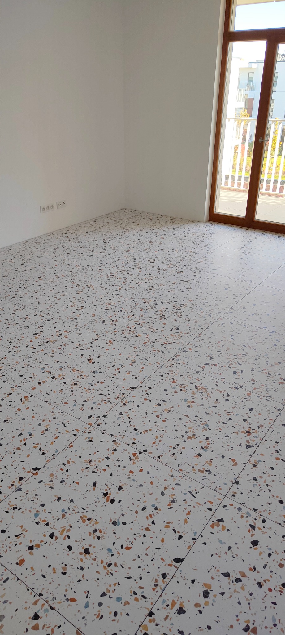 Nowoczesna posadzka z płytek lastryko w jasnym wnętrzu, widok na ścianę z gniazdkami i okno balkonowe z widokiem na budynek.