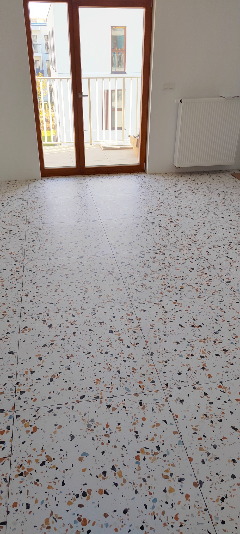 Biała posadzka terazzo z kolorowymi, drobnymi kamyczkami, widoczna z perspektywy podłogi, z oknem balkonowym i grzejnikiem w tle.