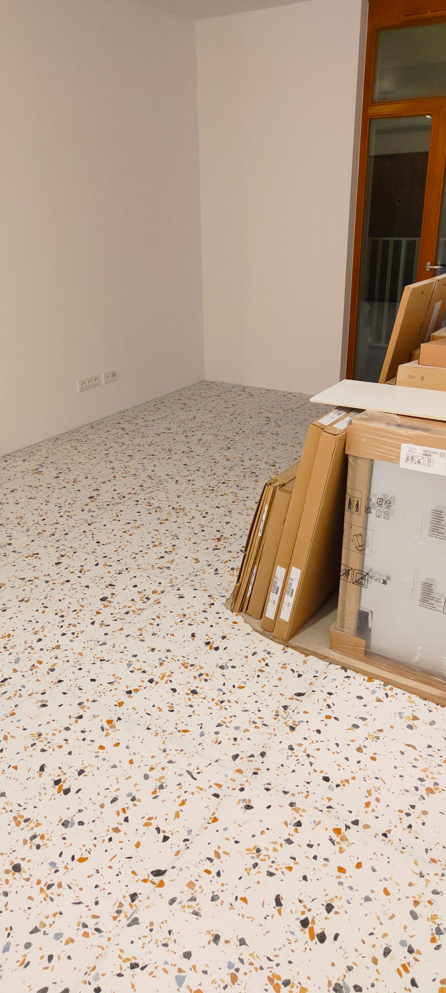 Nowe płytki terazzo na podłodze w pustym pokoju, kartony z płytkami oparte o ścianę, widoczne gniazdka elektryczne.