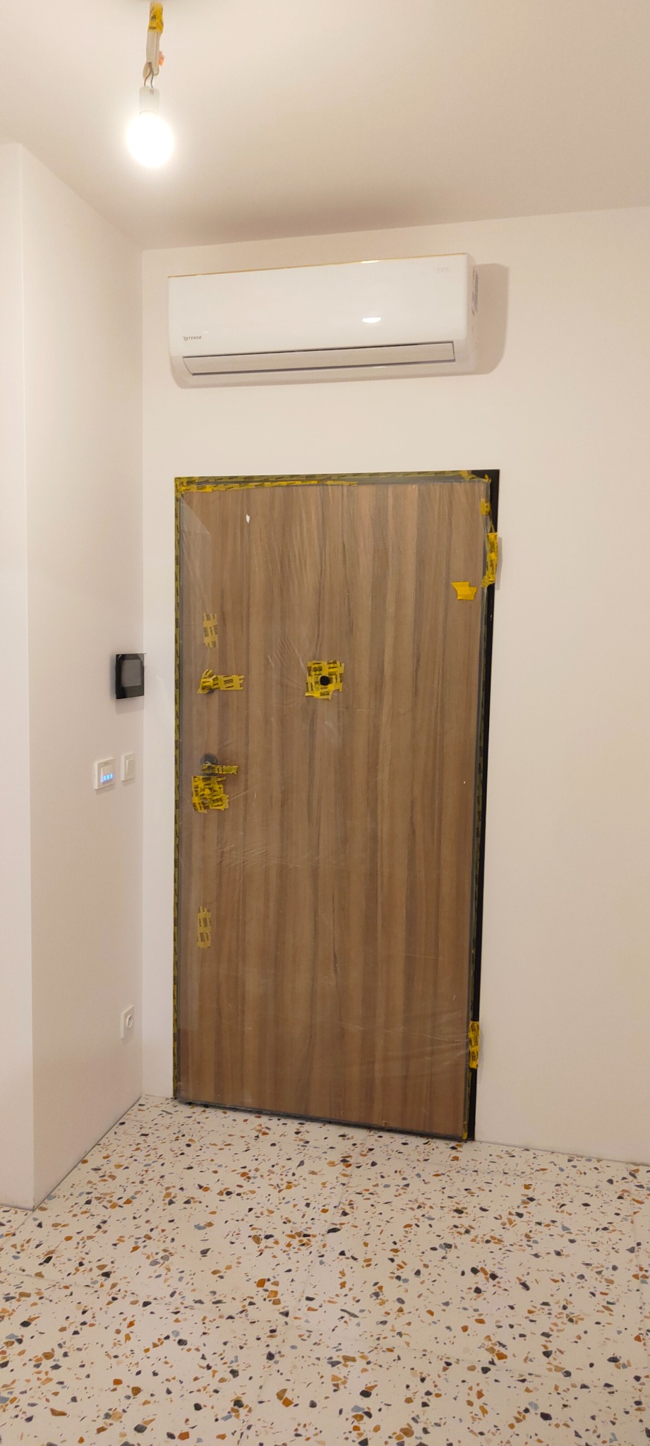 Nowe drzwi wewnętrzne z okleiną drewnopodobną, zabezpieczone folią i taśmą malarską podczas prac wykończeniowych, widoczny fragment podłogi z lastryko i klimatyzator.