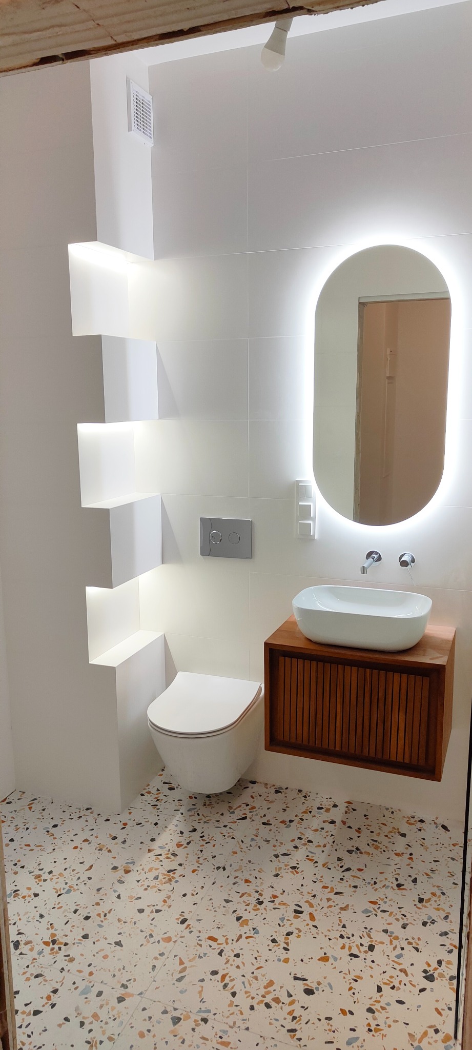 Nowoczesna łazienka z podwieszaną toaletą, umywalką nablatową na drewnianej szafce i owalnym lustrem z podświetleniem LED, wykończona białymi płytkami i posadzką typu lastryko.