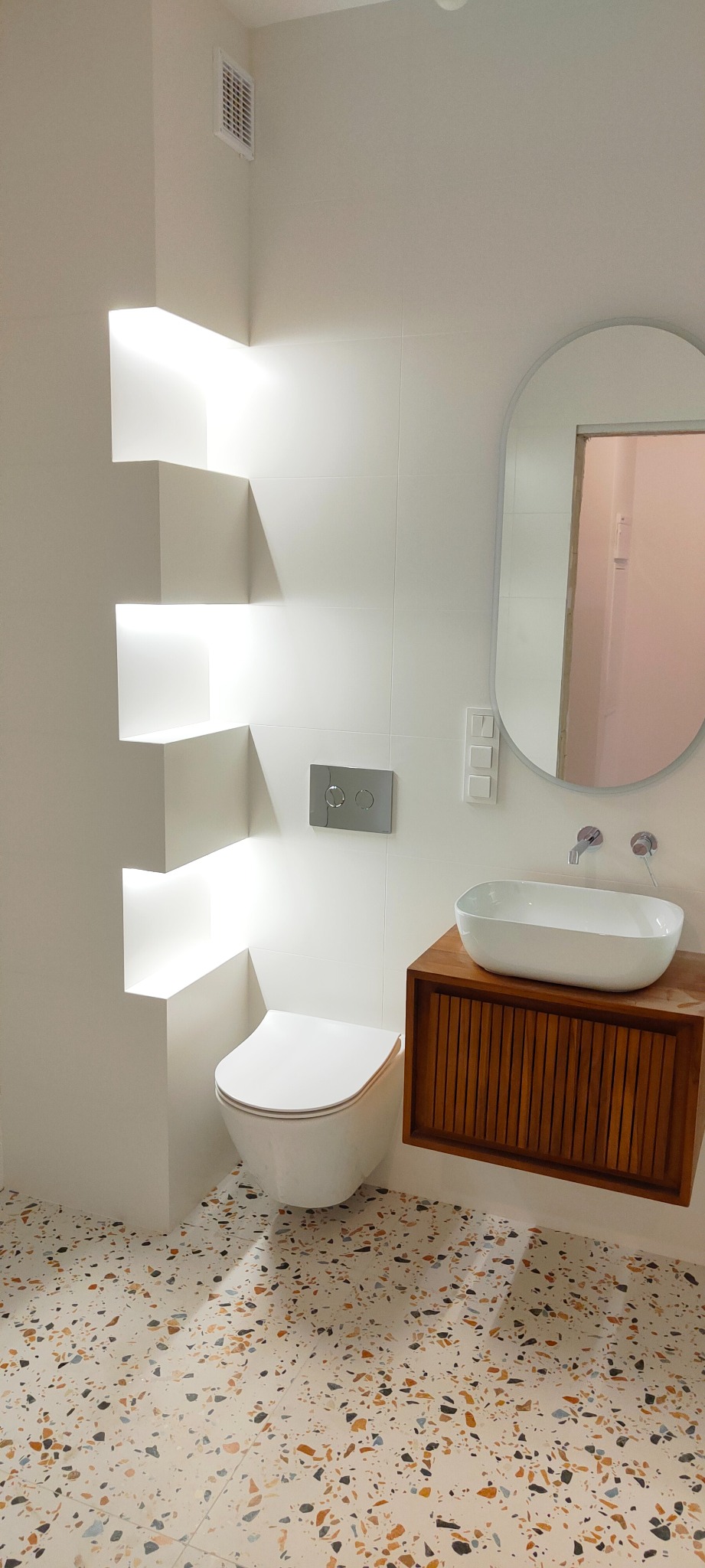 Nowoczesna łazienka z białymi płytkami, wiszącą miską WC, umywalką nablatową na drewnianej szafce, lustrem owalnym i podświetlanymi półkami w ścianie, wykończona podłogą lastryko.