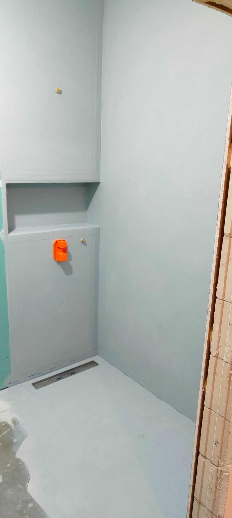 Przygotowana do wykończenia kabina prysznicowa z odpływem liniowym i półką wnękową, ściany pokryte szarą powłoką hydroizolacyjną, widoczne elementy instalacji wodnej i fragment ściany z cegieł.