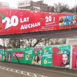 Długi baner reklamowy Auchan w Łodzi, umieszczony wzdłuż ulicy, z motywami urodzinowymi i informacją o loterii, widoczny z perspektywy przejeżdżających samochodów.
