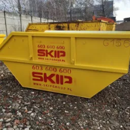 Żółty, trapezoidalny kontener z numerem G156 i logo firmy SKIP ustawiony na gruzowisku, z innymi kontenerami w tle.