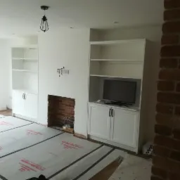 Białe meble na wymiar w salonie w trakcie montażu, zabezpieczona podłoga, widoczny fragment ściany z cegły i przygotowane miejsce na kominek.