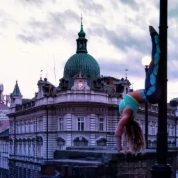 Kobieta w stroju sportowym wykonuje st&oacute;jkę przy latarni na tle zabytkowego budynku Poczty Polskiej z zegarem na fasadzie.