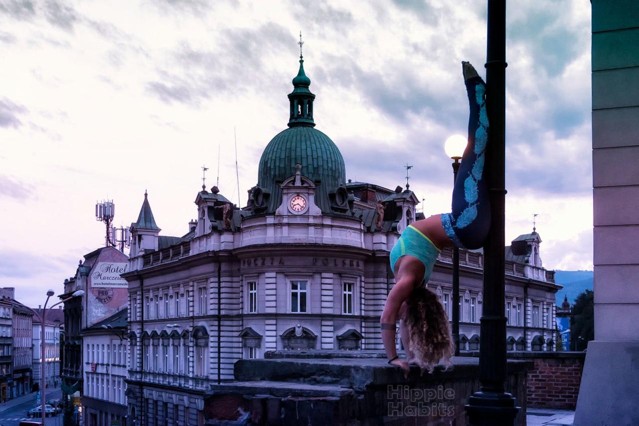 Kobieta w stroju sportowym wykonuje stójkę przy latarni na tle zabytkowego budynku Poczty Polskiej z zegarem na fasadzie.