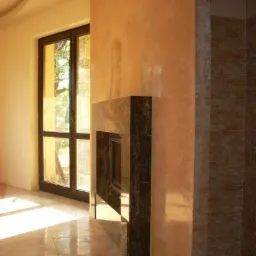 Wnętrze z wykończonym kominkiem obłożonym ciemnym kamieniem, ściana pokryta dekoracyjnym tynkiem w odcieniach beżu i brązu, fragment ściany wykończony cegłą dekoracyjną, światło słoneczne wpadające...