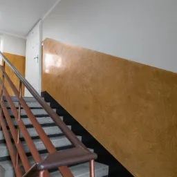 Klatka schodowa w budynku wielorodzinnym po remoncie, ściany pomalowane na kolor miodowy z efektem dekoracyjnym, szare betonowe schody z brązową metalową balustradą, kwiaty w doniczce na parapecie...