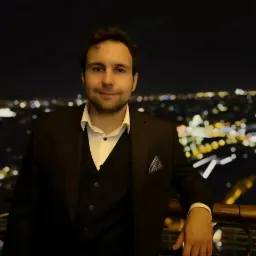 Mężczyzna w garniturze na tle nocnej panoramy miasta z rozmytymi światłami, oparty o balustradę. W tle widoczna wieża Eiffla.