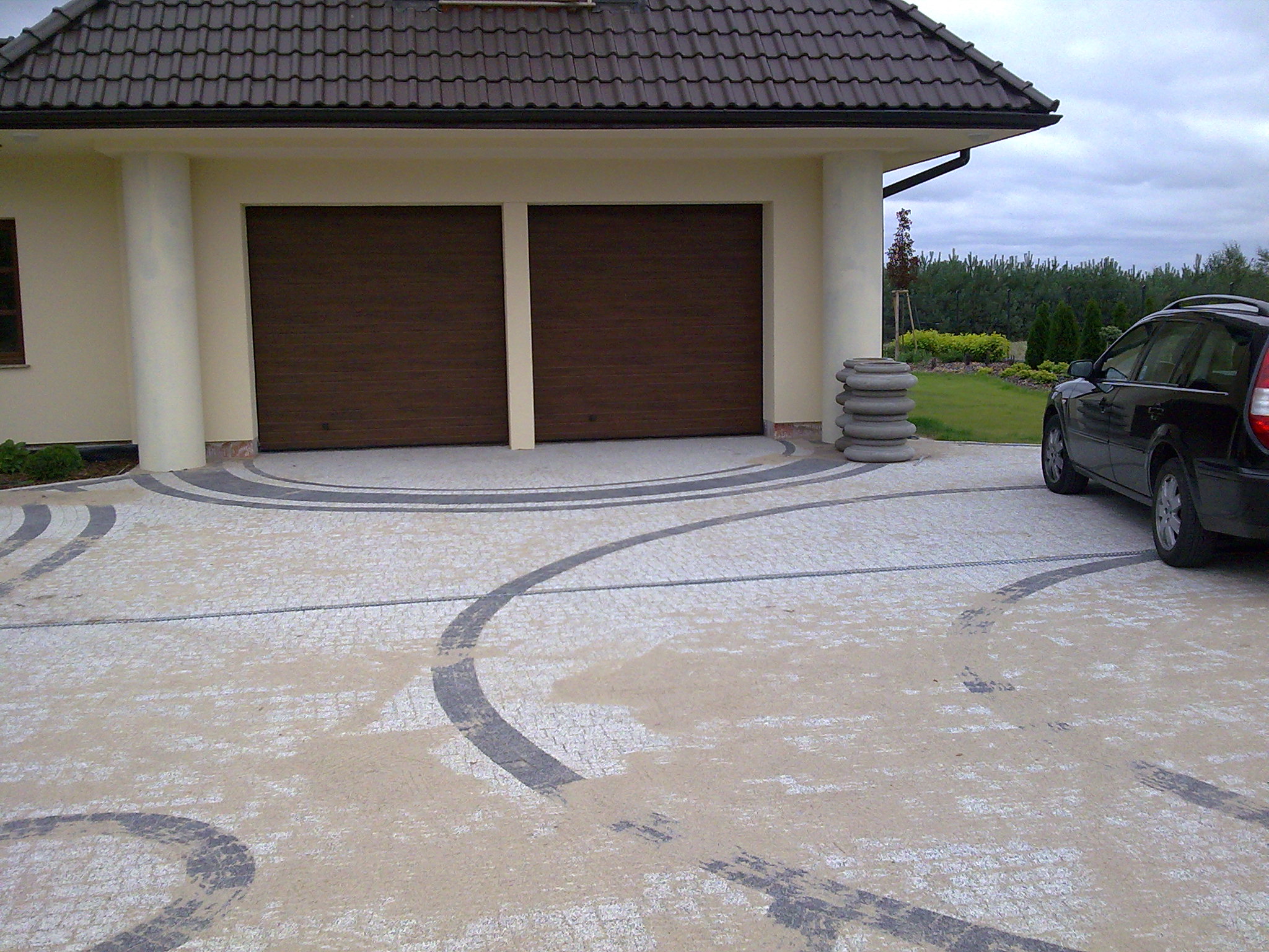 Podjazd do garażu wyłożony jasną, drobną kostką z ciemnoszarymi, okrągłymi wzorami; widoczne ślady opon, sterta betonowych kręgów i fragment czarnego samochodu.