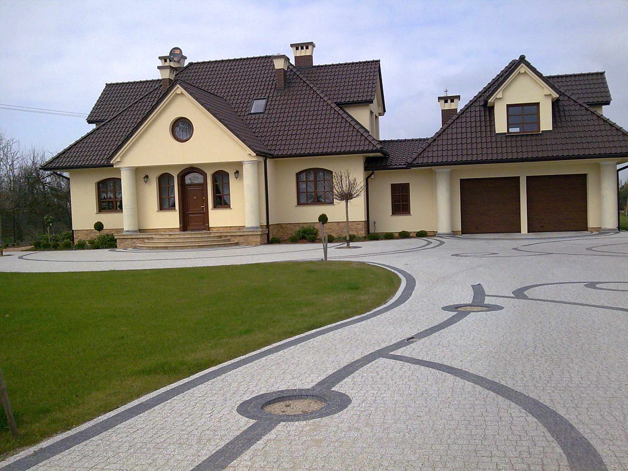 Elegancki dom jednorodzinny z beżową elewacją i brązowym dachem, otoczony trawnikiem i podjazdem wyłożonym jasną kostką z ciemnoszarymi akcentami.