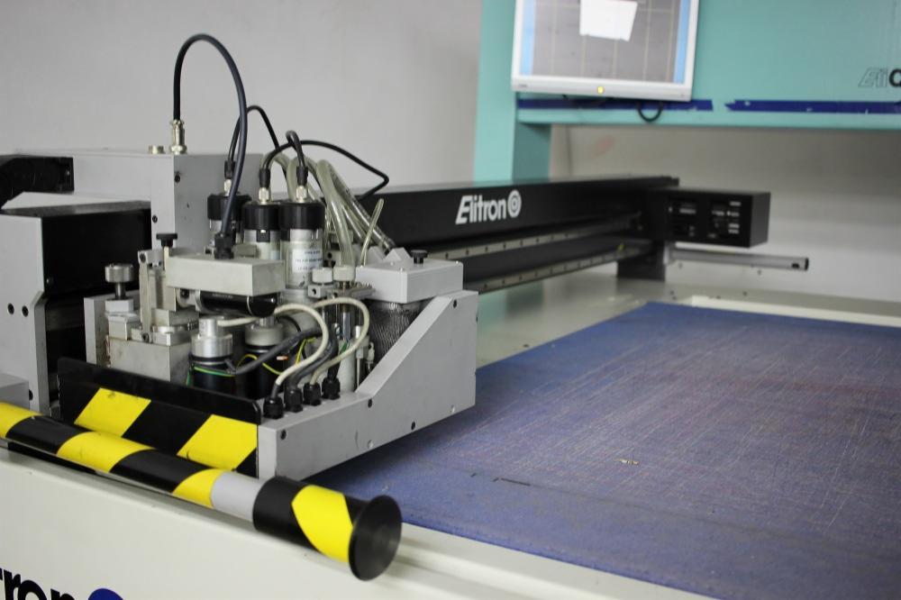 Automatyczna maszyna do cięcia materiałów tekstylnych marki Elitron, widoczna niebieska mata z drobnymi śladami cięć, czarno-żółta osłona zabezpieczająca z przodu.