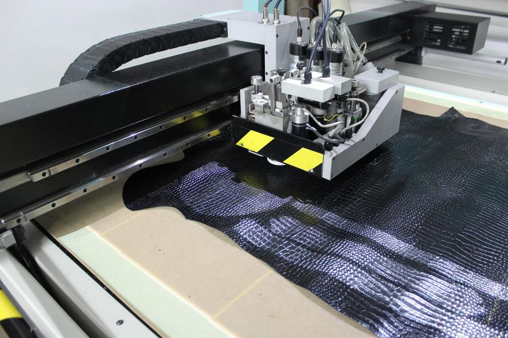 Automatyczna krojownica tnąca czarną skórę o fakturze krokodylej, widoczne elementy mechaniczne i przewody urządzenia.