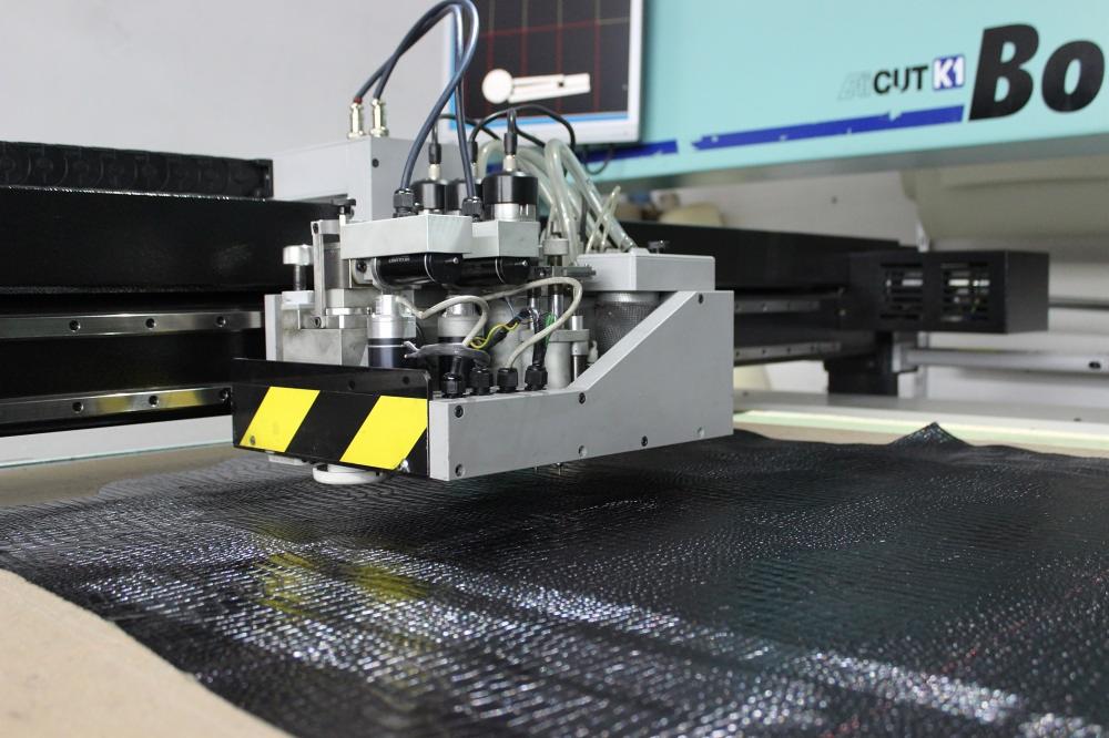 Automatyczna maszyna tnąca precyzyjnie wycina wzór z czarnego materiału tekstylnego o wyraźnej fakturze, widoczna głowica tnąca z żółto-czarnymi pasami ostrzegawczymi.