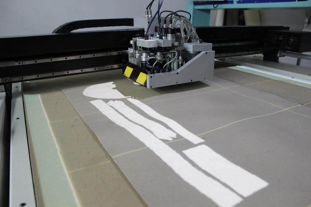 Automatyczna maszyna tnąca precyzyjnie wycina elementy z arkusza materiału, pozostawiając białe ślady po cięciu na szarym podłożu z zaznaczoną siatką.