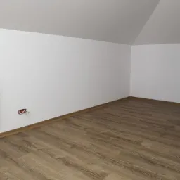 Puste, jasne pomieszczenie na poddaszu w Markach z drewnianą podłogą i gładkimi, białymi ścianami. Widoczny otwór instalacyjny w ścianie.