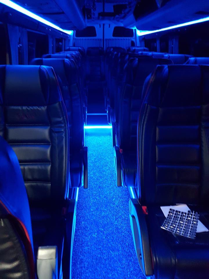 Wnętrze autobusu z czarnymi skórzanymi siedzeniami oświetlone niebieskim światłem LED na podłodze i suficie, widoczne numery siedzeń na oparciu.