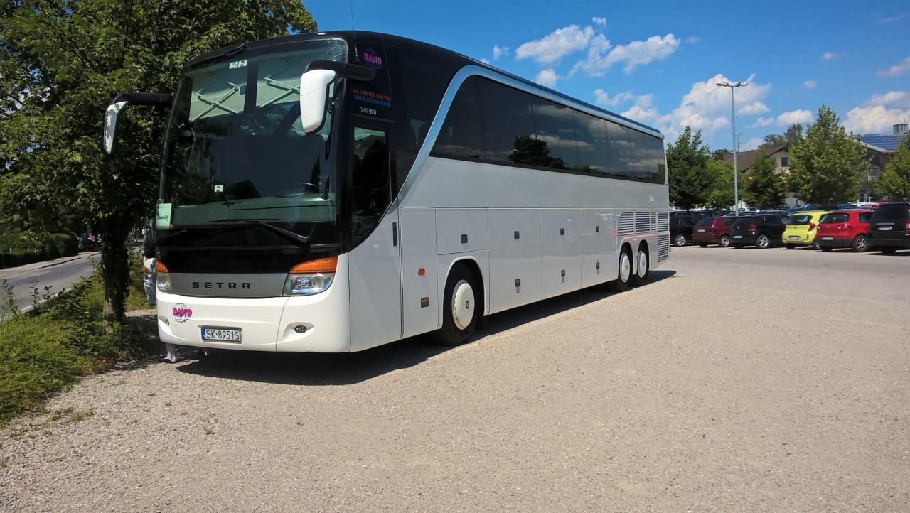 Biało-szary autokar marki Setra parkuje na żwirowym parkingu z widocznymi drzewami i innymi samochodami w tle, pod błękitnym niebem z lekkimi chmurami.