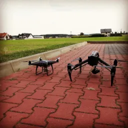 Dwa drony na czerwonej kostce brukowej, w tle zielone pole i domy; jeden dron mniejszy z białymi końcówkami śmigieł, drugi większy z kamerą pod spodem.