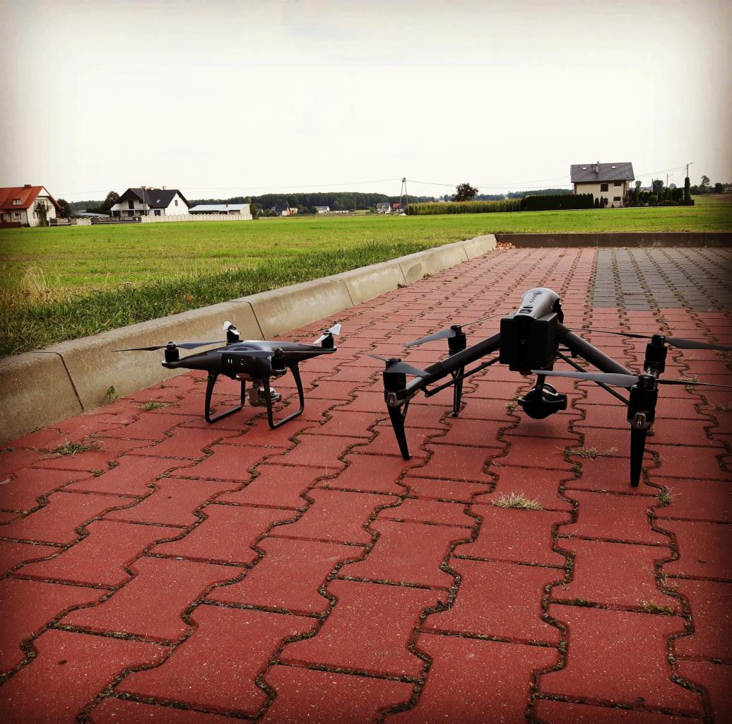 Dwa drony na czerwonej kostce brukowej, w tle zielone pole i domy; jeden dron mniejszy z białymi końcówkami śmigieł, drugi większy z kamerą pod spodem.
