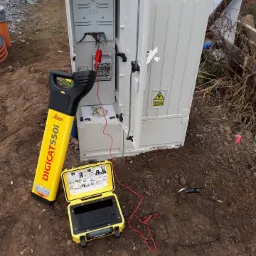 Żółty lokalizator kabli Leica DIGICAT 550i, otwarta skrzynka narzędziowa i podłączone przewody pomiarowe w ziemi przy otwartej szafie elektrycznej z widocznymi elementami instalacji.