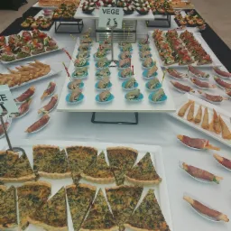 Bogato zastawiony st&oacute;ł z r&oacute;żnorodnymi przekąskami, w tym tartaletki ze szpinakiem, roladki z szynki parmeńskiej, mini tortille i koreczki, z tabliczkami informacyjnymi 'Vege'.