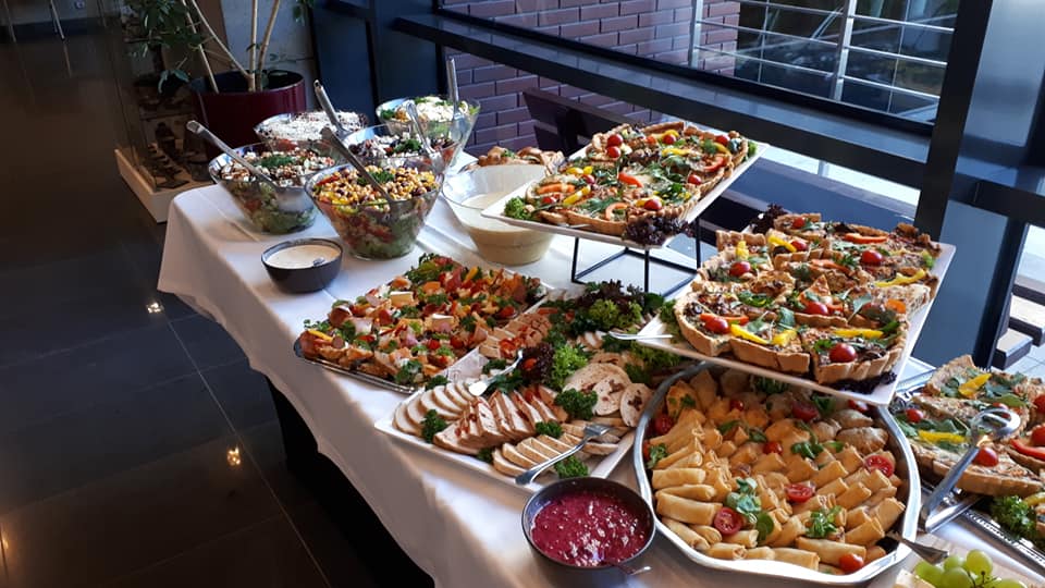 Stół cateringowy z różnorodnymi potrawami: sałatki w szklanych misach, tarty z warzywami na paterach, wędliny w plastrach, roladki z ciasta francuskiego, sos żurawinowy w rondelku, całość na białym...