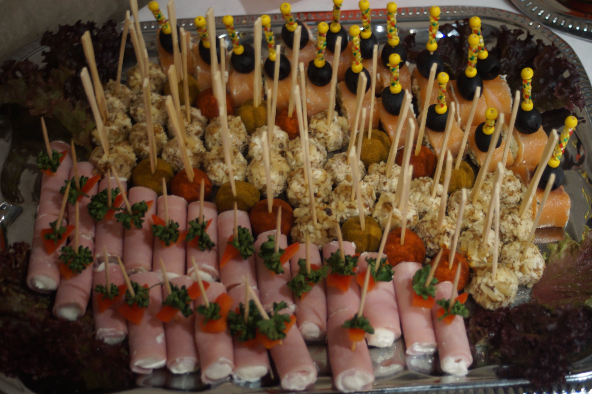 Srebrna patera z różnorodnymi przekąskami na drewnianych wykałaczkach: roladki z szynki z farszem, kulki serowe obtoczone w orzechach, koreczki z łososia i oliwek, dekorowane sałatą.