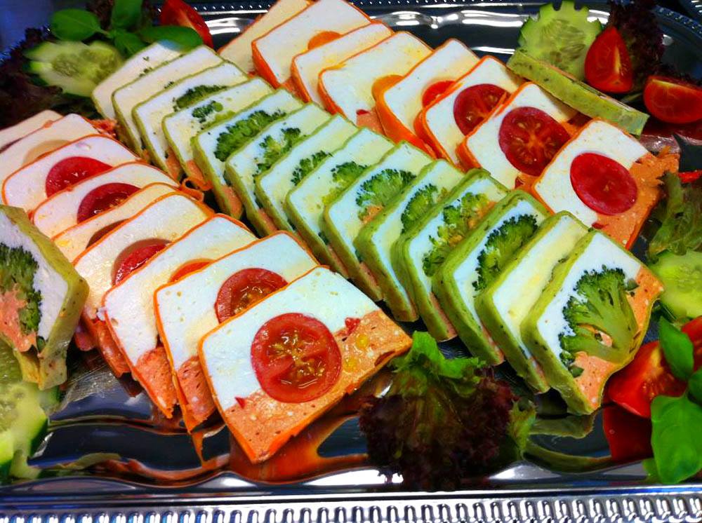 Elegancko ułożone na srebrnej tacy przekąski: trójkątne kanapki z nadzieniem warzywnym (brokuły, pomidory) i białym serem, udekorowane świeżymi ogórkami i sałatą.
