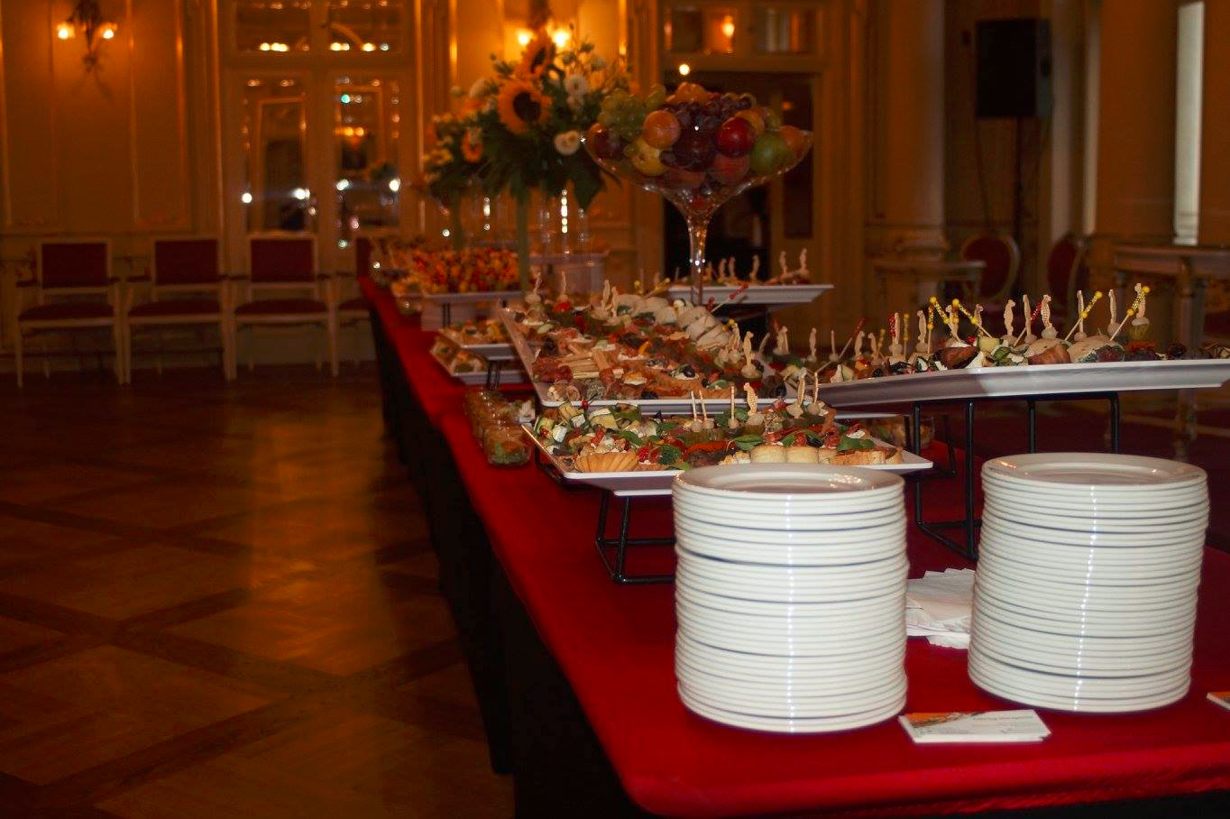 Długi stół przykryty czerwonym obrusem, zastawiony przekąskami na białych paterach, owocami w szklanej wazie i stosami białych talerzy, w tle eleganckie wnętrze sali balowej z lustrami i oświetleniem