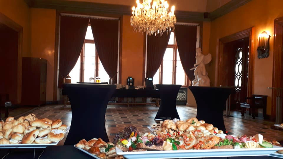 Eleganckie wnętrze z przygotowanym stołem cateringowym: kanapki, ciastka, ekspresy do kawy, żyrandol kryształowy, brązowe zasłony, posadzka w szachownicę.